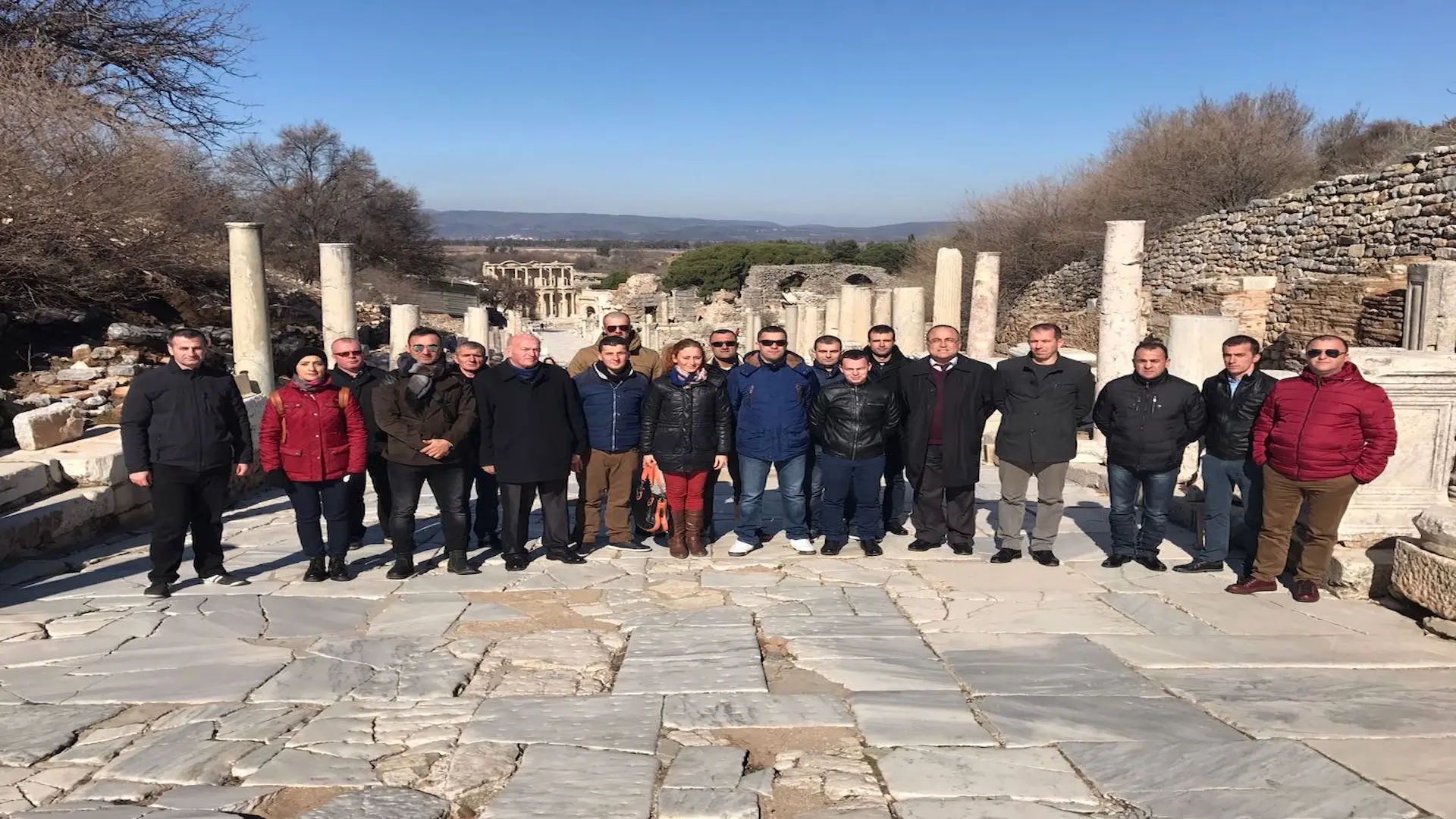 A large group of tourists posing on the ancient Marble Road with the Library of Celsus in the distance.