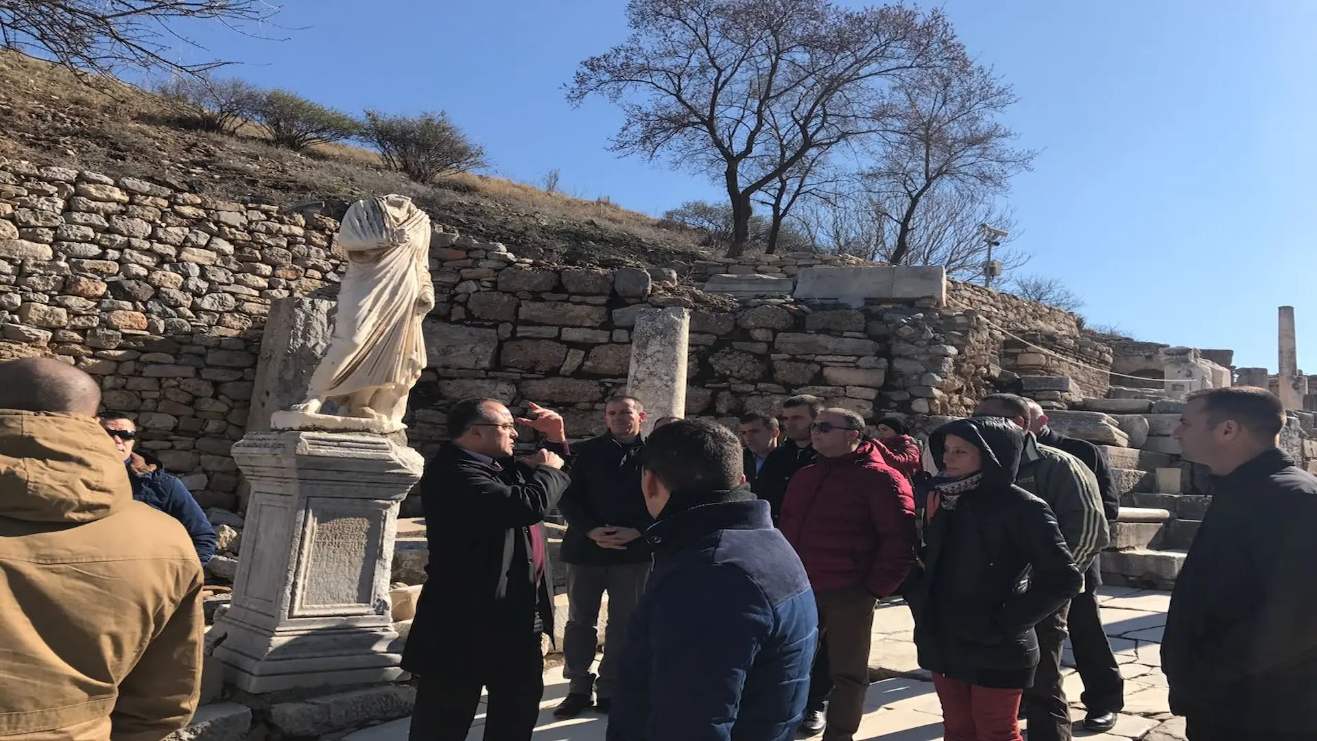 A group of tourists listening to a guide's lecture in front of a headless Roman statue in Ephesus.
